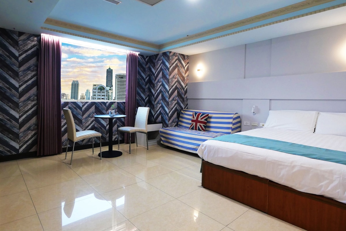 Habitación de hotel con cama, sofá, una mesita y una silla. Tiene una ventana con cortinas, y el diseño es moderno con vista a la ciudad.