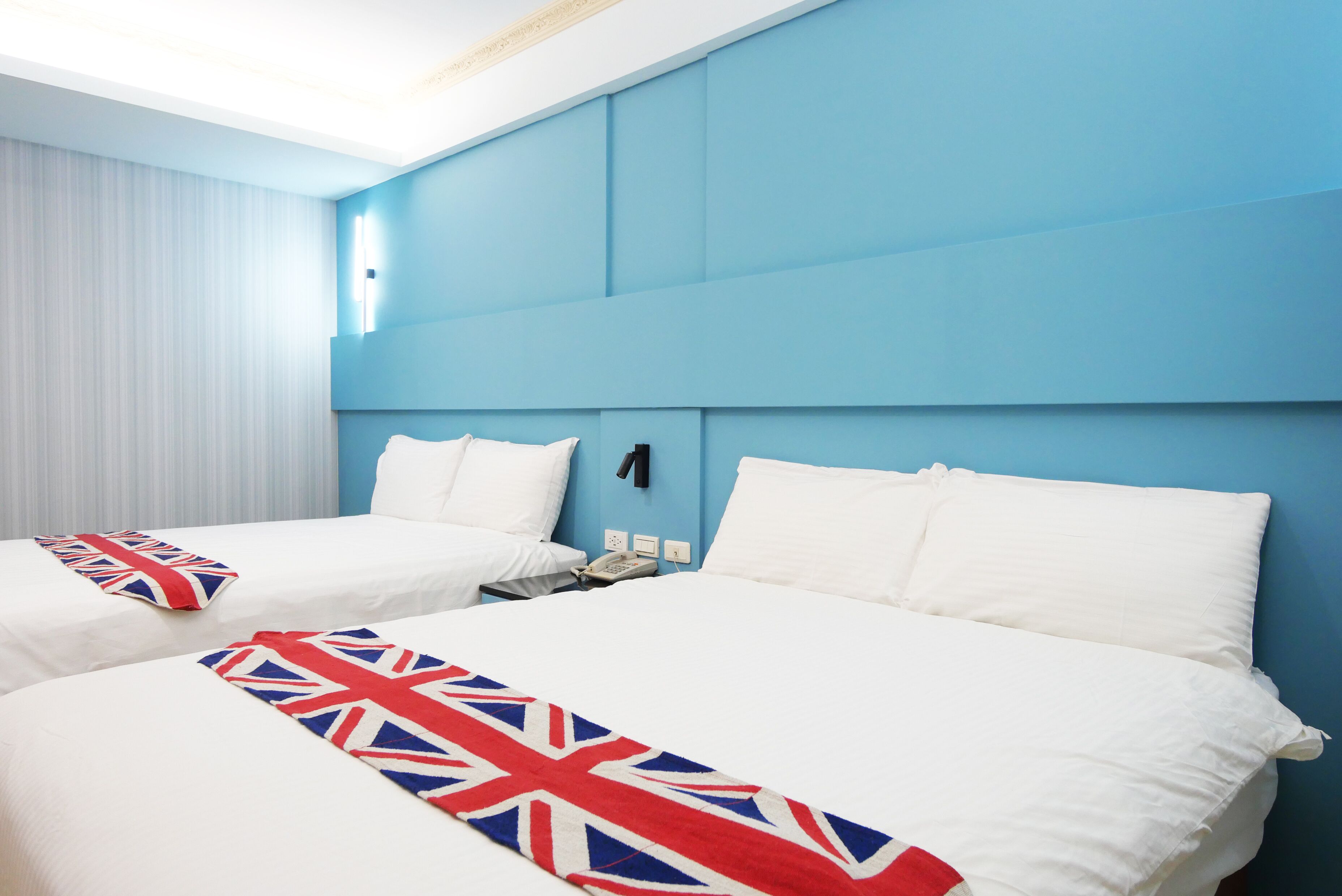 A hotel room with two beds, each with white bedding and a Union Jack blanket. The room has blue walls and a white ceiling.