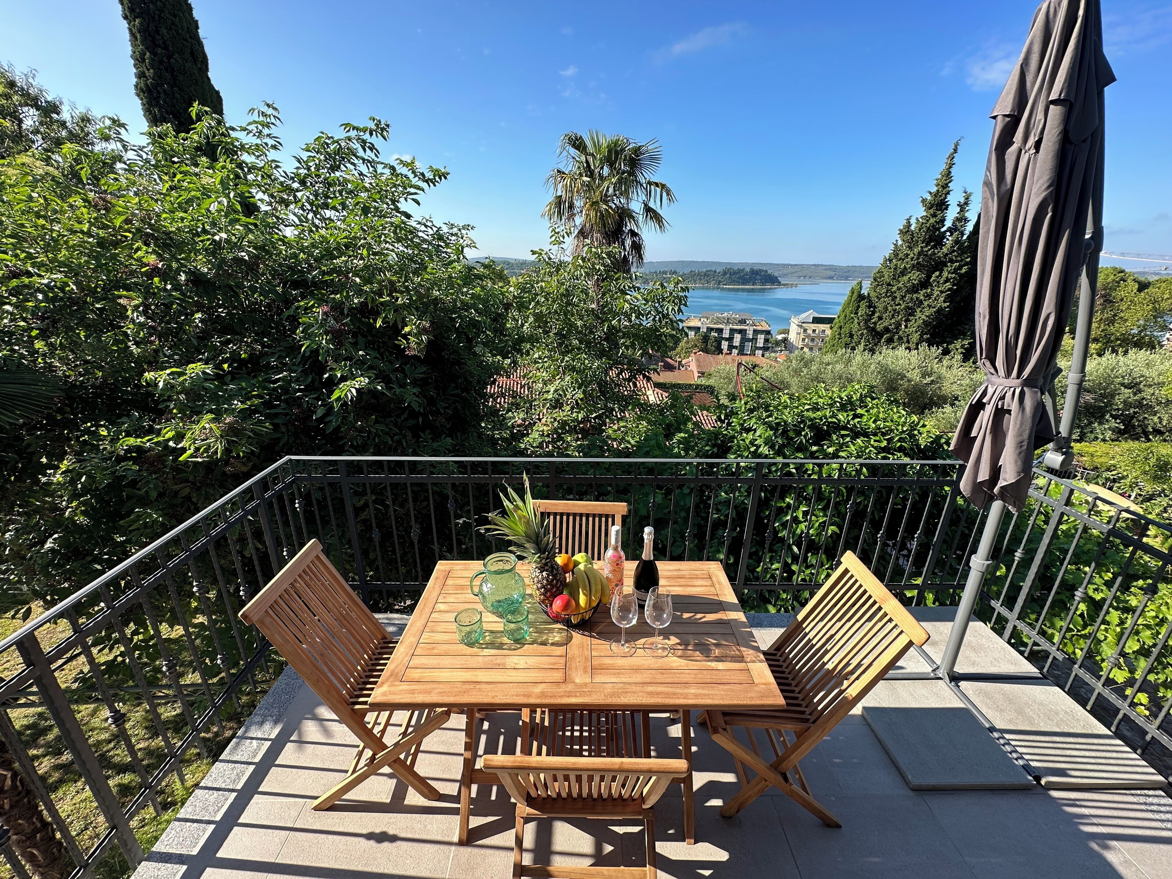 A wooden table with chairs, a bottle of champagne, and a fruit bowl on a balcony with a view of the sea and buildings.