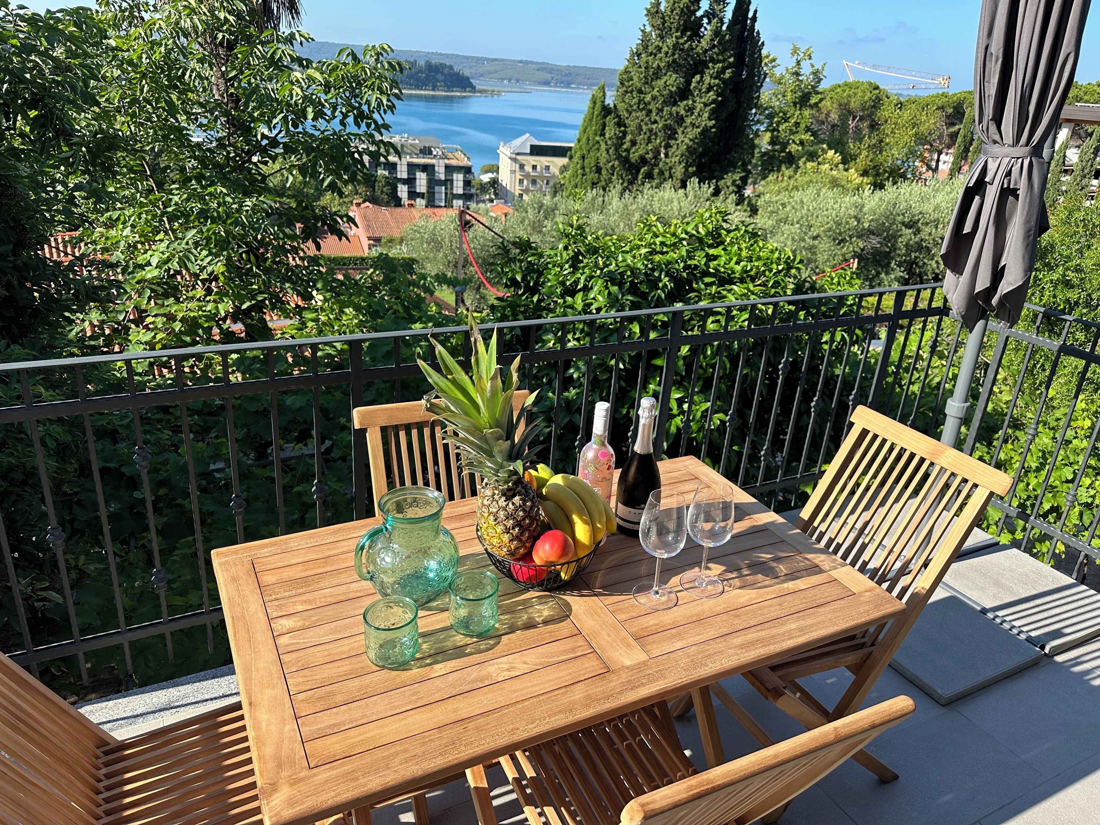Een houten tafel met een fles, twee glazen, een ananas en een schaal met fruit op een balkon met uitzicht op een mooi waterlandschap en gebouwen.