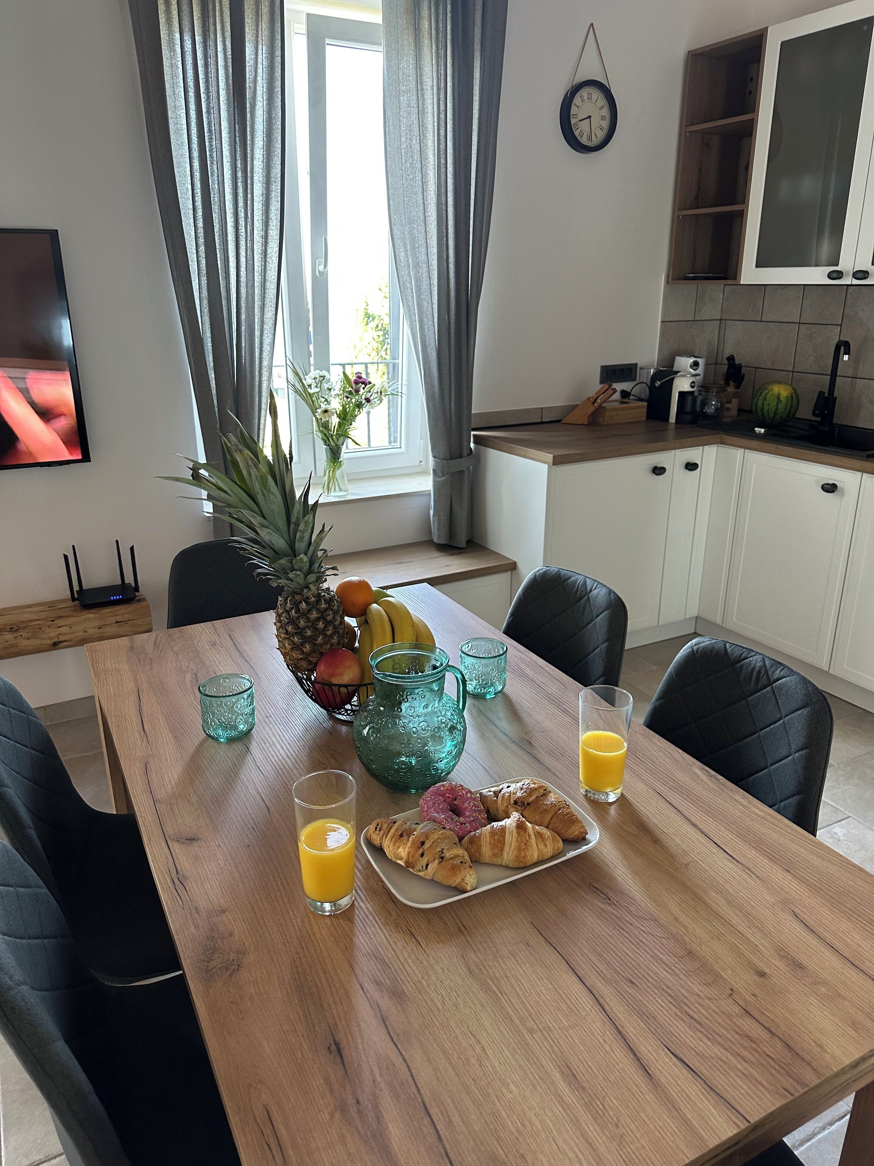 A dining area with a wooden table set for breakfast, including croissants, juice, and a fruit bowl.