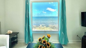 A room with a large window overlooking a beach, a small table with a vase of sunflowers, and a black TV stand. - East End Paradise Villas 4Bd3.5B-Private Beach-On the Water-w/ Sea Level Studio (East End)