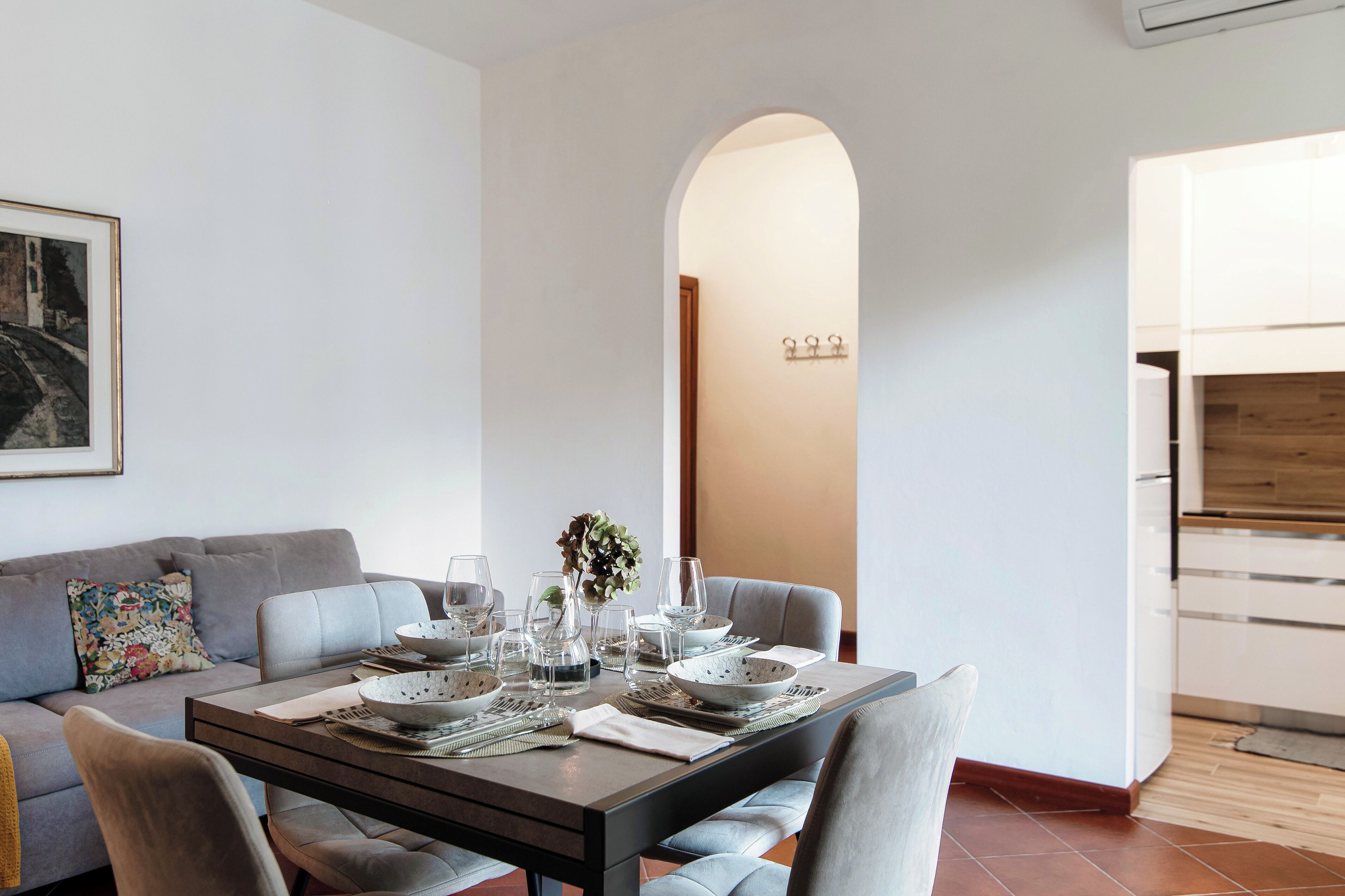 A dining area with a table set for four, a sofa with a colorful pillow, and a kitchen in the background.