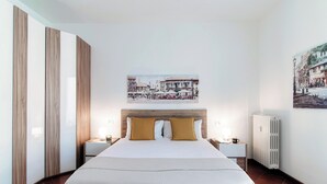 A hotel room with a large bed, wooden wardrobe, white walls, and a cityscape painting.