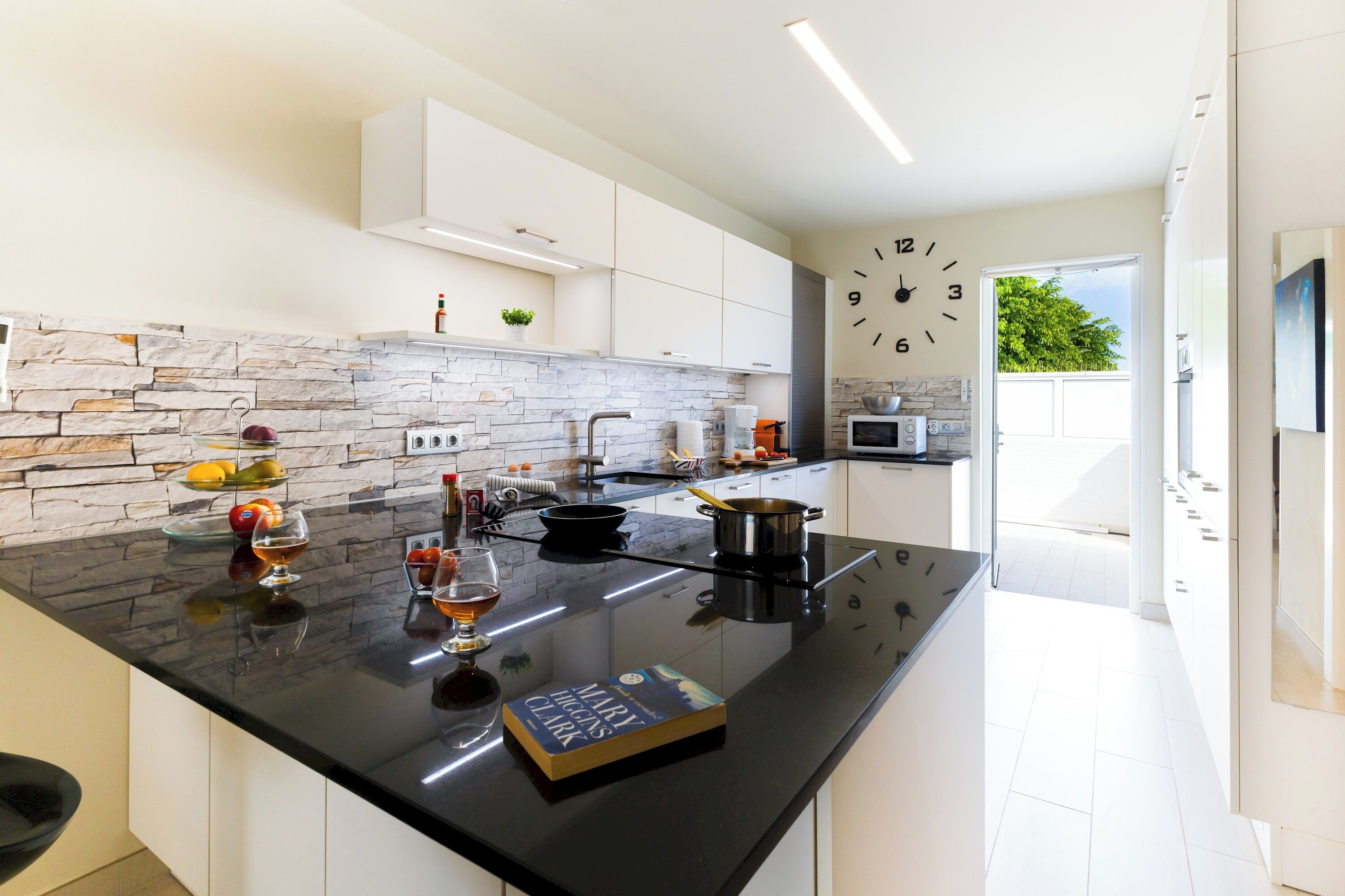 A modern kitchen with a black countertop, white cabinets, and a stone backsplash. There is a clock on the wall and a window providing natural light.
