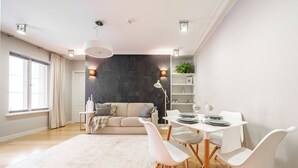 A modern living room with a sofa, dining table, and chairs. There is a large window with curtains, a plant on a shelf, and a white rug on the floor. - Charming 1-bedroom apartment with WIFI in Old Tallinn (Tallinn)