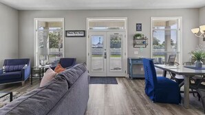 A living room with a grey sofa, a blue armchair, a dining table with chairs, and a view of the street through a glass door.