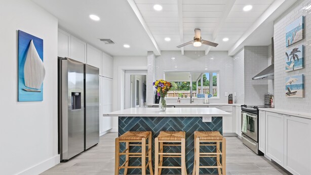 A modern kitchen with white cabinetry, a stainless steel refrigerator, a central island with stools, and a ceiling fan. - 237 | Heated Pool | Putting | Outdoor Living | FreeBee Zone | by DelrayNightly (Delray Beach)