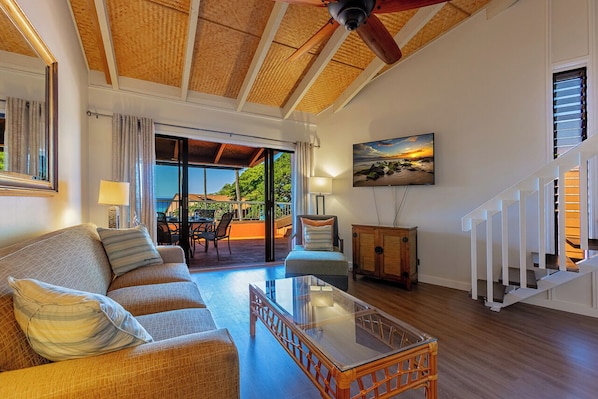 A spacious living area with a high ceiling, a glass coffee table, a sofa, and a view of the outdoors through large sliding doors. - Oceanfront 2BR condo with lanai and resort pool in Lahaina summer deals (Lahaina)