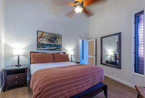 A bedroom with a bed, two bedside tables with lamps, a ceiling fan, a large mirror, and a window with blinds. - Oceanfront 2BR condo with lanai and resort pool in Lahaina summer deals (Lahaina)