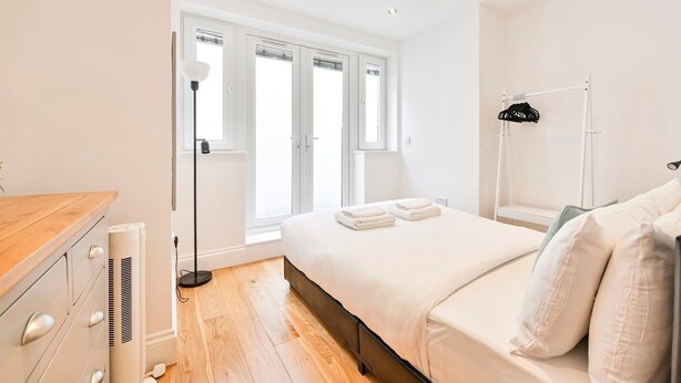 A modern bedroom with a wooden dresser, a bed with white linens, a standing lamp, and a window with blinds.