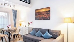 A modern living room with a grey sofa, blue cushions, a dining table with chairs, a white dresser, and a floor lamp.