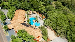 An aerial view of a resort with a swimming pool, a thatched-roof building, and lush greenery surrounding a body of water.