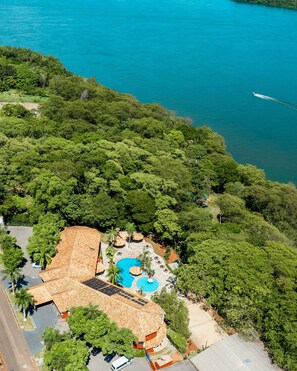 An aerial view of a resort with a swimming pool, a thatched-roof building, and lush greenery surrounding a body of water.