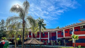 Une piscine dotée d’une terrasse en pierre, entourée de palmiers et bordée d’un bâtiment rouge avec de nombreuses fenêtres et des antennes paraboliques.