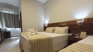 A hotel room with two beds, a wooden headboard, a bedside table with a phone and a notepad, and a window with curtains.