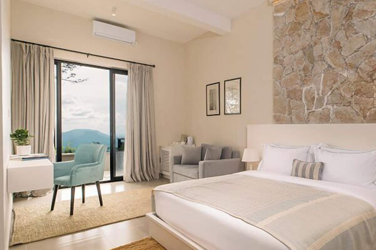 A bedroom with a large bed, a desk with a chair, a sofa, and a stone wall.