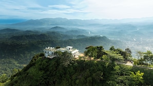 A modern building with a flat roof, surrounded by dense forest and multiple umbrella structures, overlooking a valley. - W15 Ambuluwawa (Gampola)