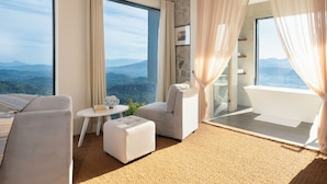 A modern hotel room with a large window offering a mountain view, a sitting area with a sofa and ottoman, a small table with a vase, and a separate bathroom with a bathtub. - W15 Ambuluwawa (Gampola)