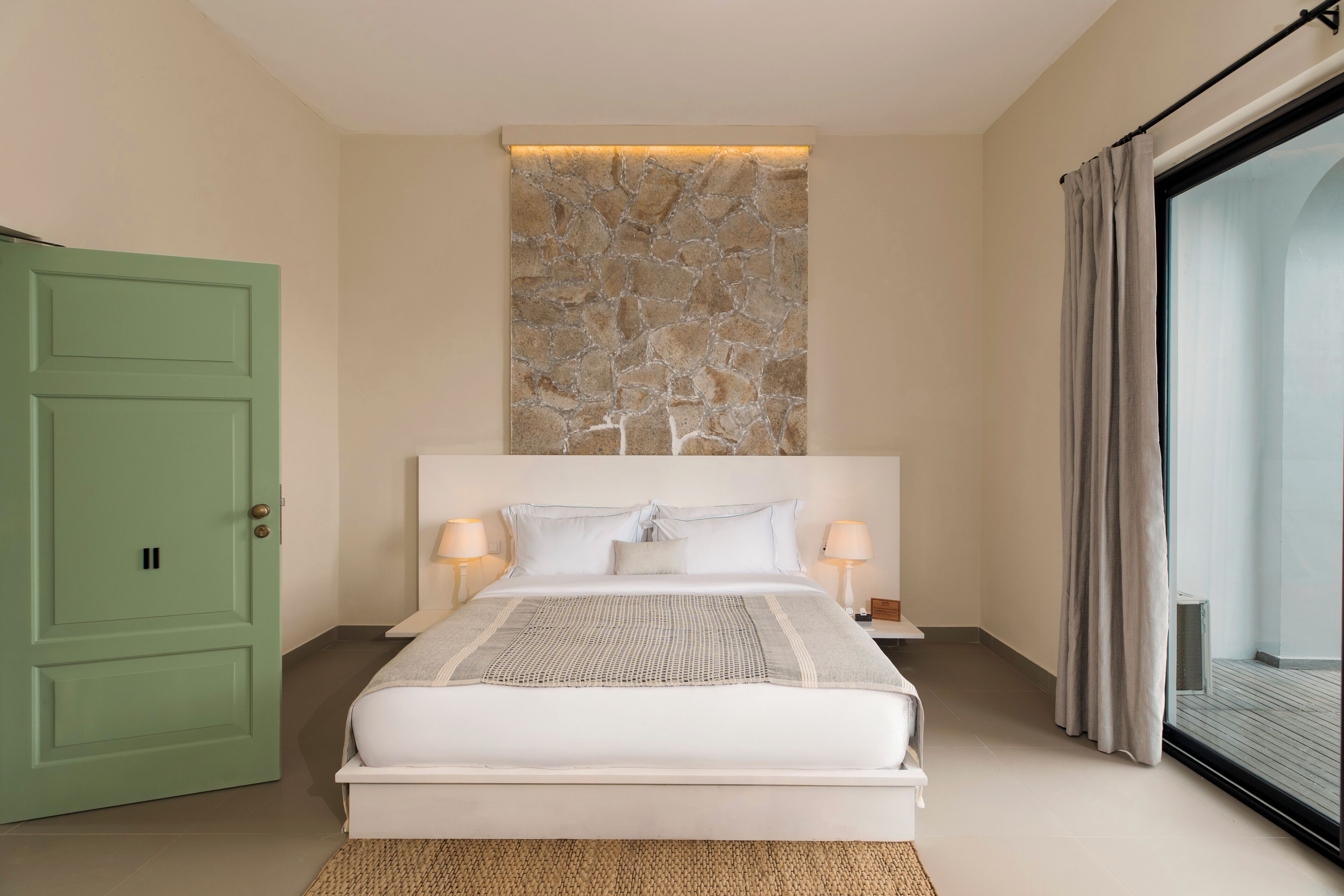 A modern bedroom with a large bed, two bedside lamps, a stone accent wall, a green door, and a view through a glass door.