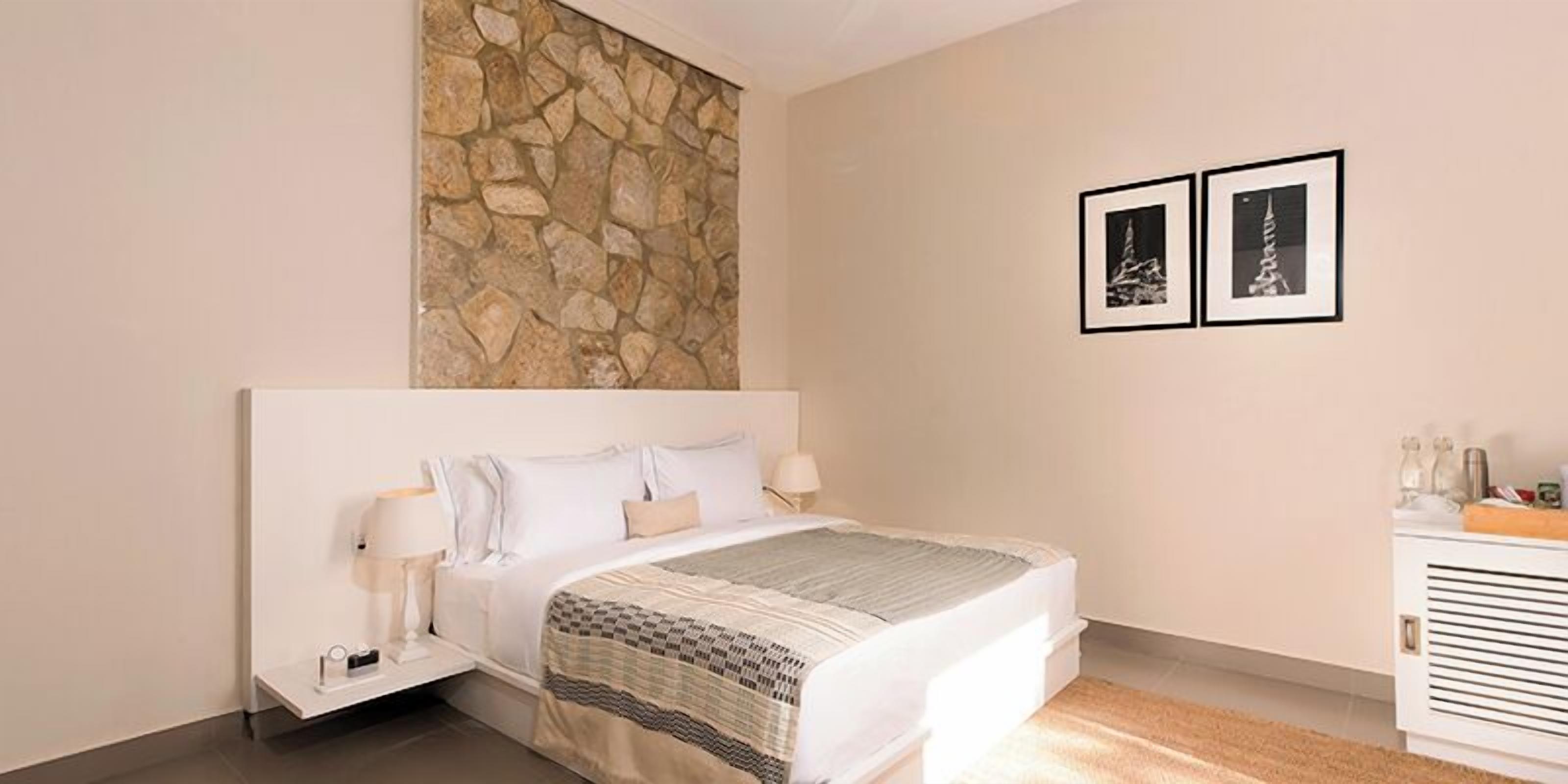 A bedroom with a stone accent wall, a bed with white linens, two framed pictures on the wall, and a bedside table with a lamp.