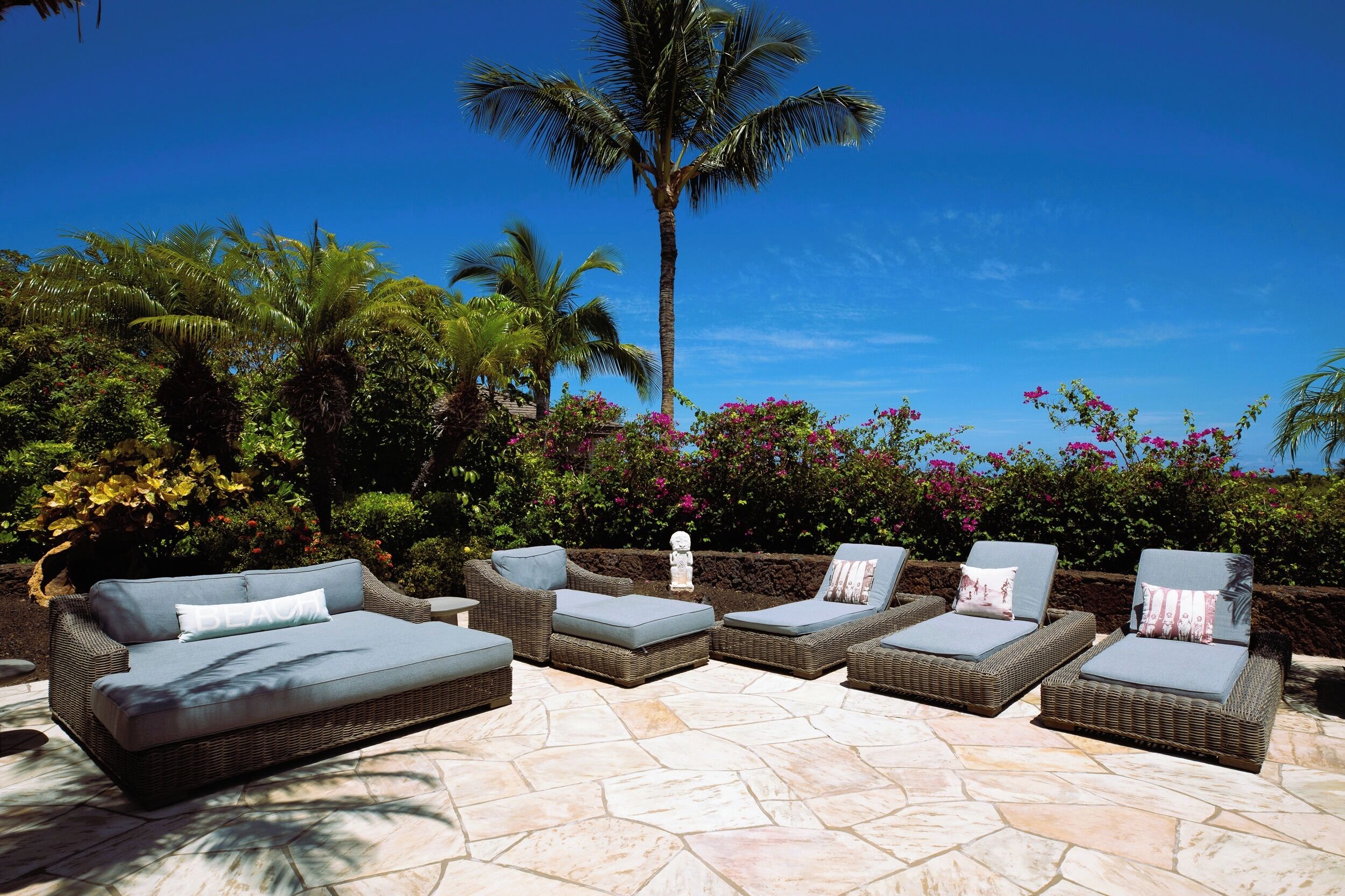 Een terras met wicker meubels, waaronder een bank en loungestoelen, gelegen op een stenen vloer met uitzicht op palmbomen en bloeiende planten.
