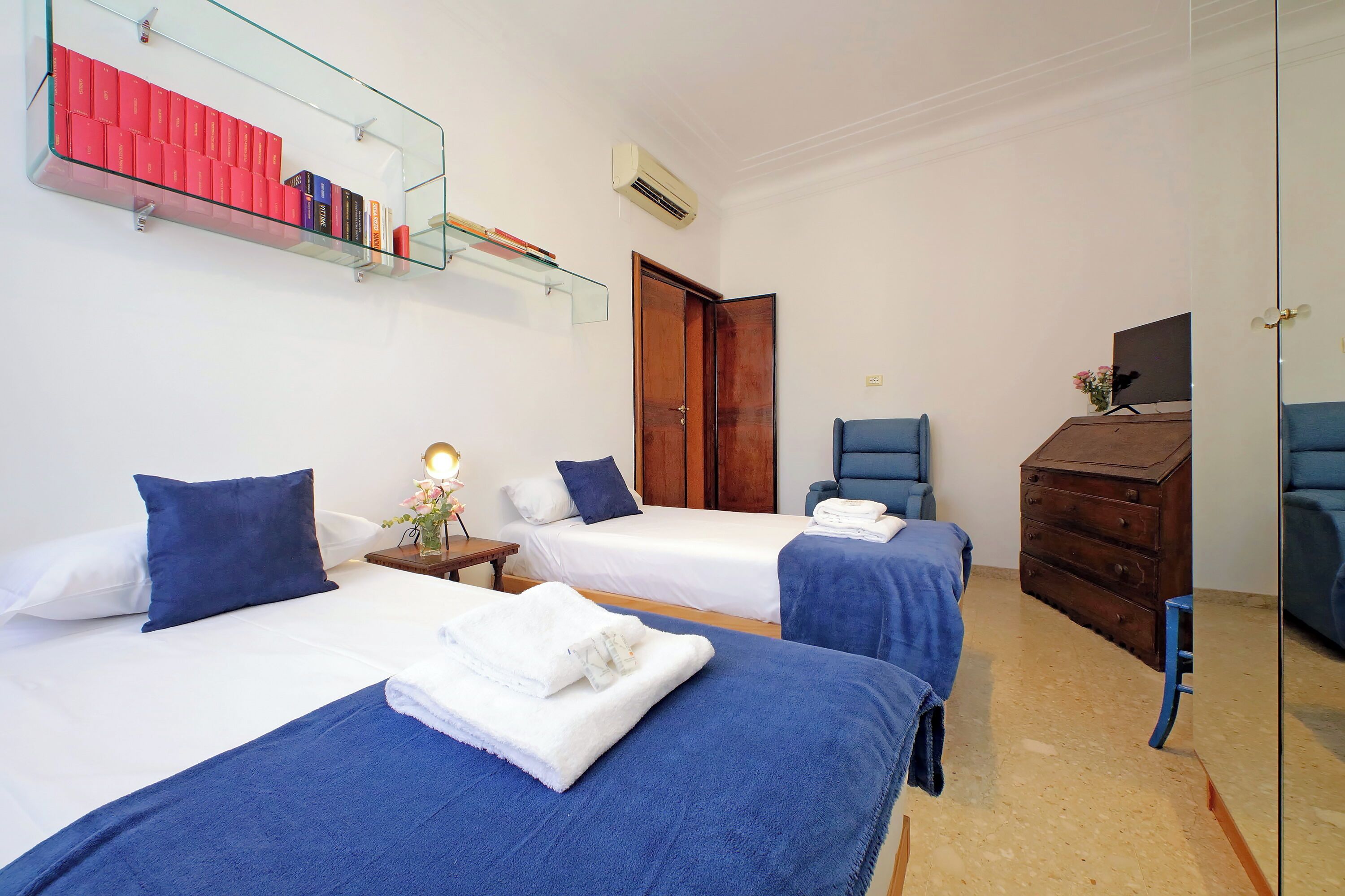 A hotel room with two beds, a wooden dresser, a TV, and a glass shelf with books.