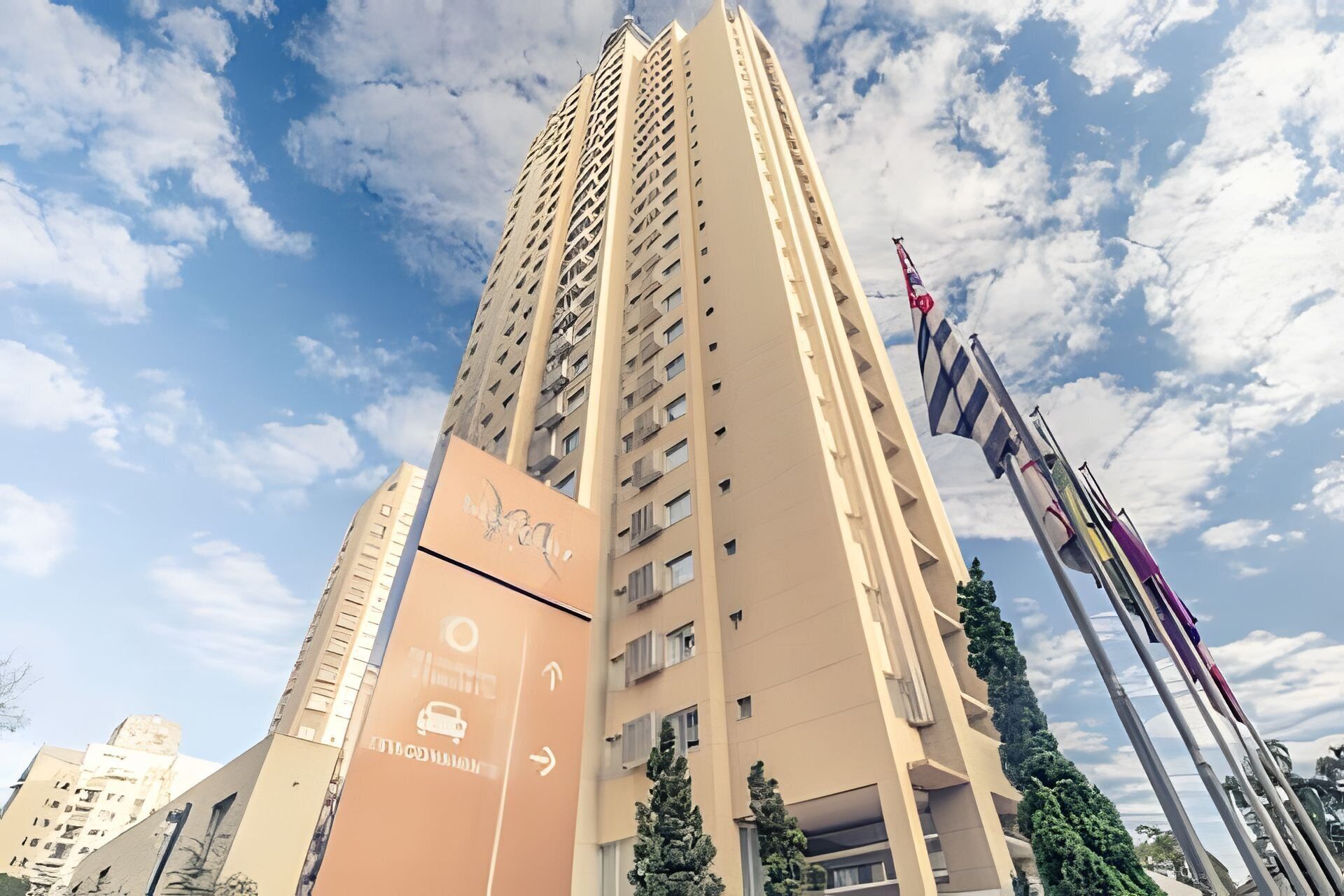 Un edificio alto y color beige con una fachada singular y varios balcones. Hay banderas en el lado derecho del edificio.