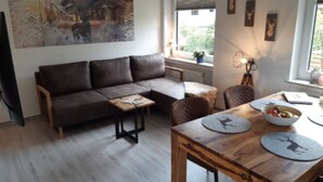 A modern living room with a brown leather sofa, a wooden coffee table, and a dining table with a bowl of fruit. There is a painting on the wall and a window with blinds. - Apartment Wurmbergblick (Braunlage)