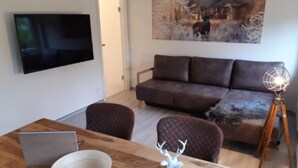 A modern living room with a flat-screen TV mounted on the wall, a brown leather sofa, a wooden dining table with a laptop and a white bowl, and a deer figurine. - Apartment Wurmbergblick (Braunlage)