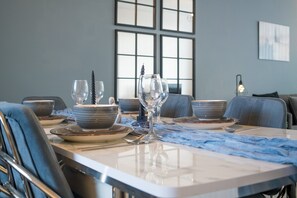 A dining table set with plates, bowls, and wine glasses, with a blue table runner and a view of a cityscape through large windows.