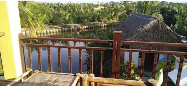 A wooden deck with a railing overlooking a calm body of water, surrounded by lush greenery and a thatched-roof structure.