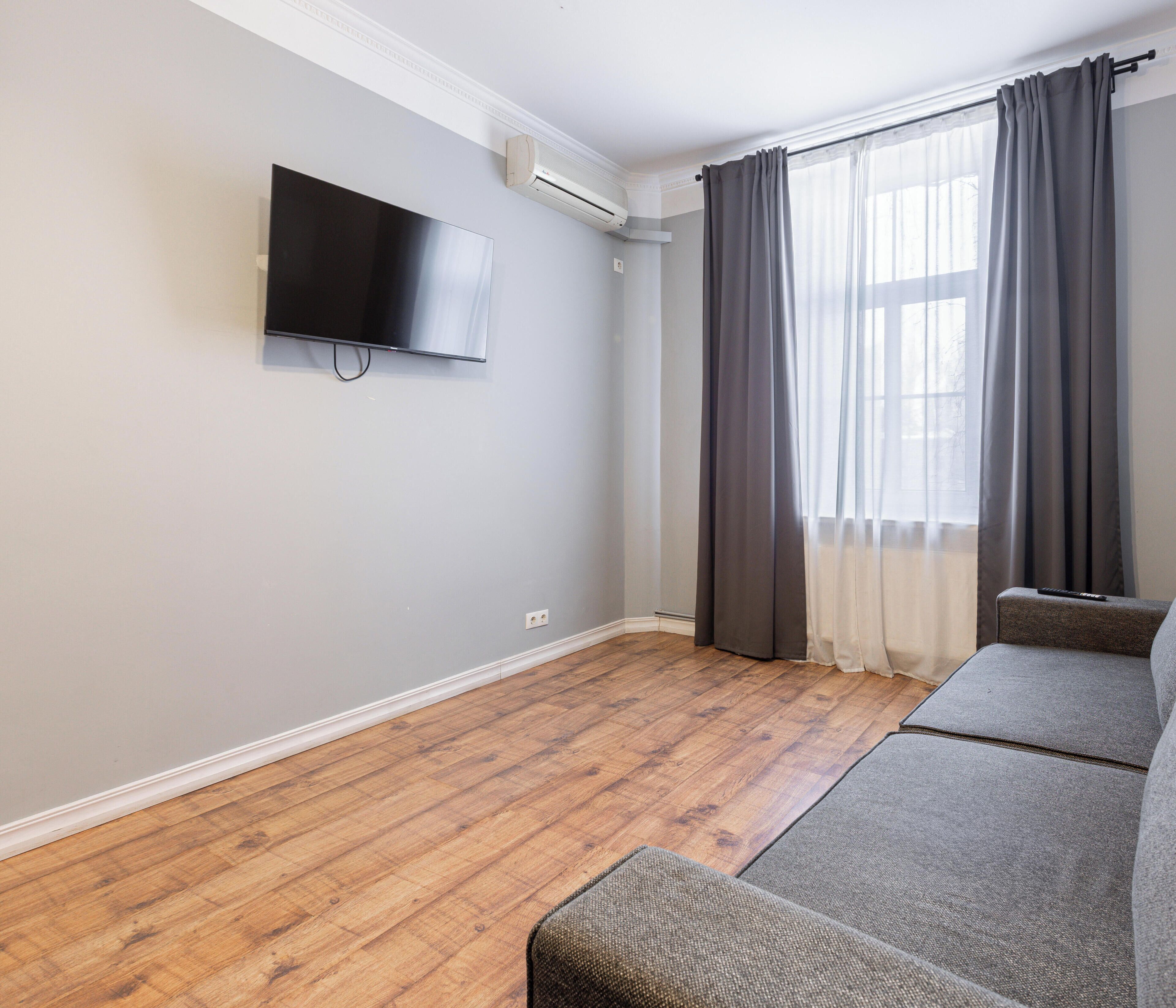 Una habitación con una televisión de pantalla plana montada en la pared, un sofá gris, una ventana con cortinas grises y un piso de madera.