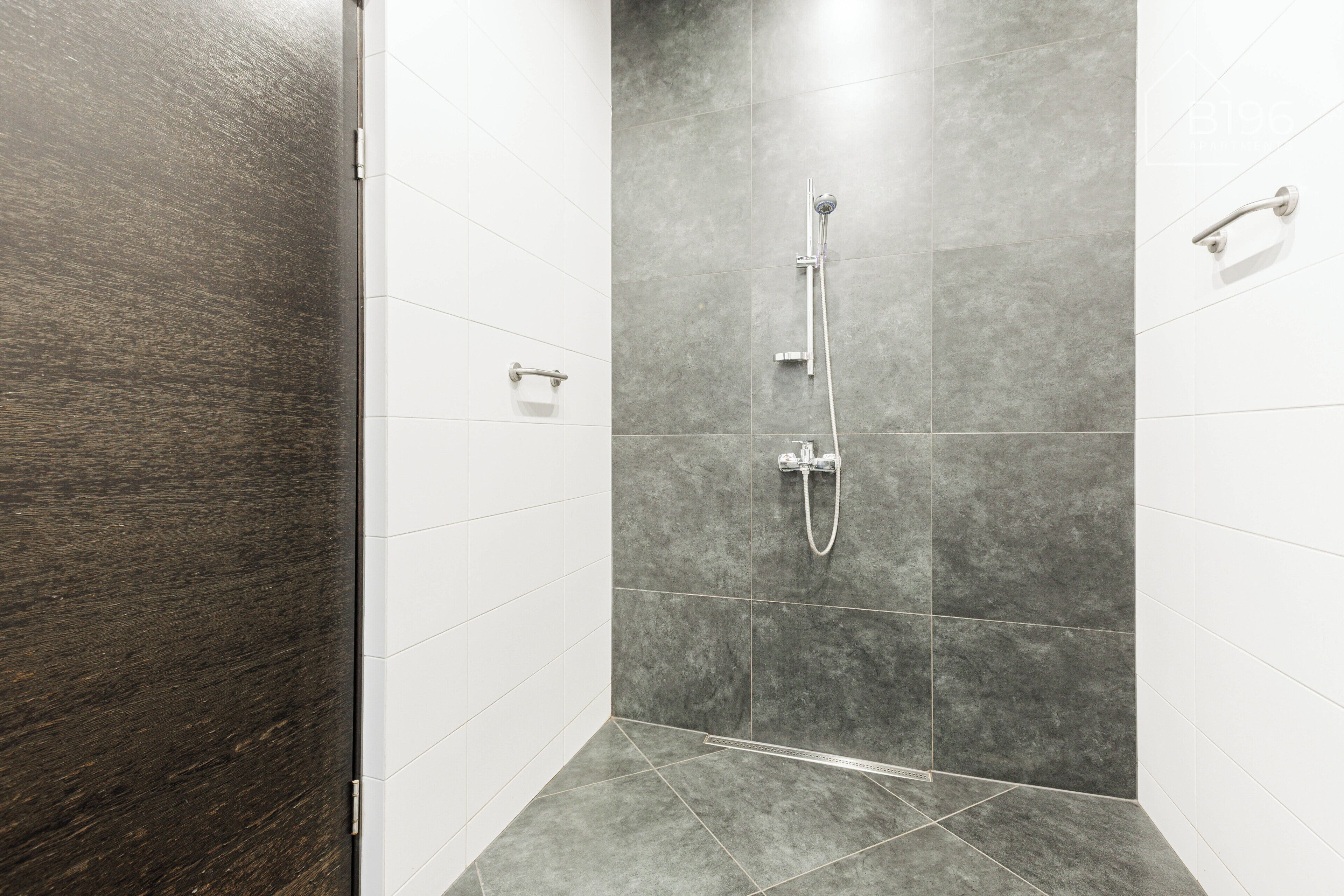 Un espace douche doté d’une porte en bois foncé, de murs recouverts de carreaux gris et d’un sol carrelé de blanc.