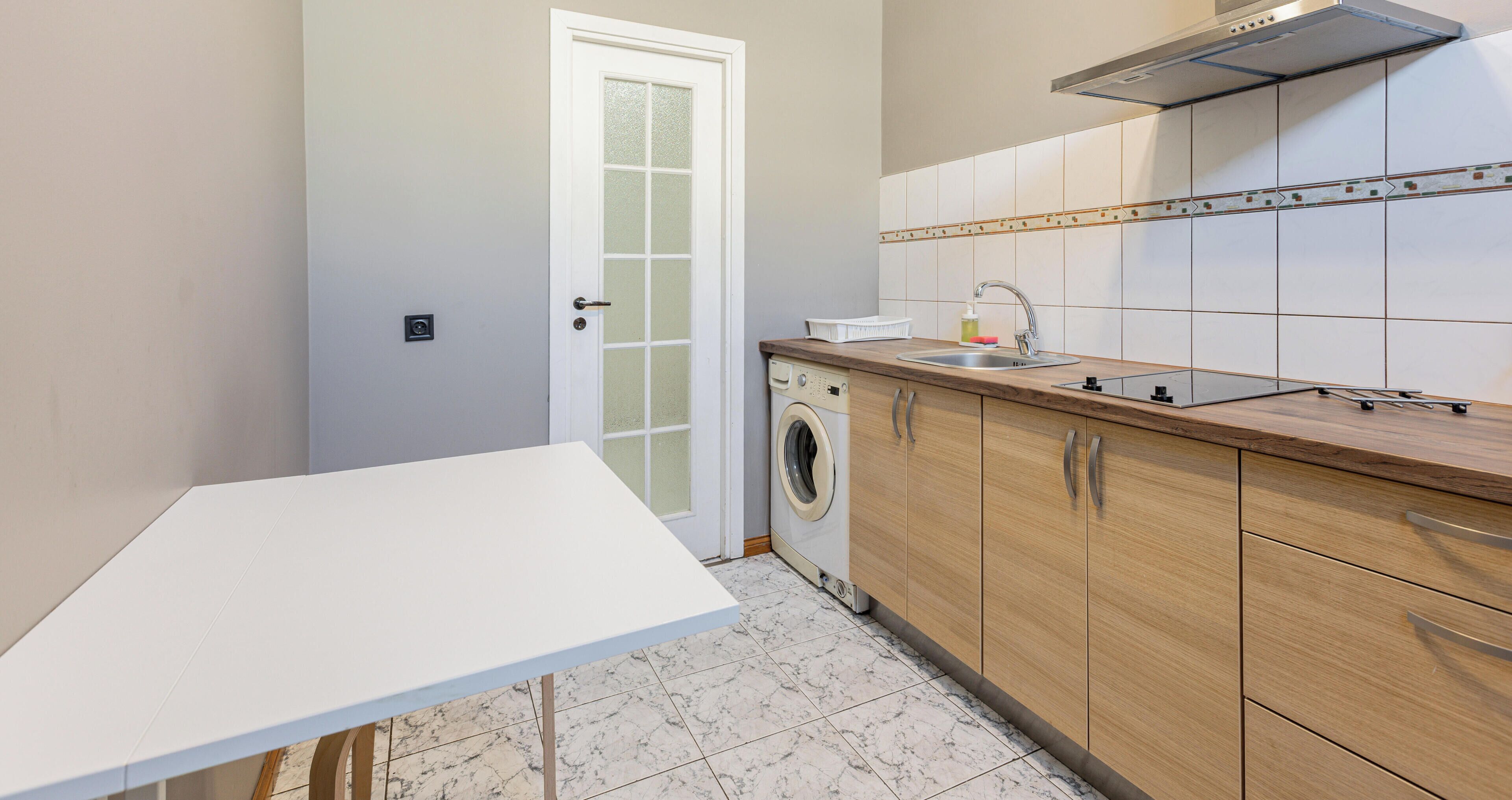 Een keuken met een wasmachine, houten kastjes, een wit aanrechtblad en een betegelde spatwand.
