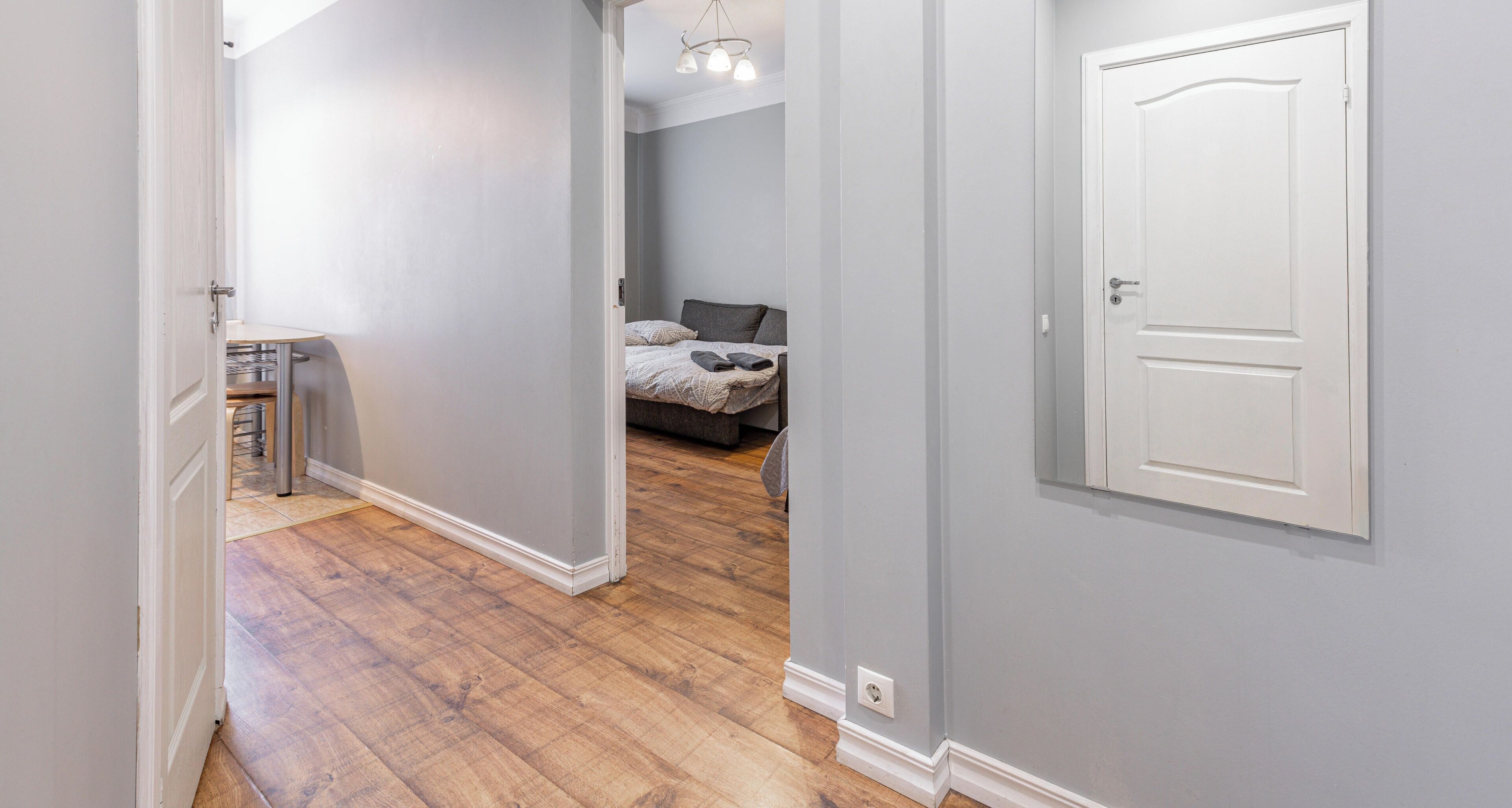 Un pasillo con piso de madera, una puerta blanca a la derecha y una cama con cabecera gris al fondo.