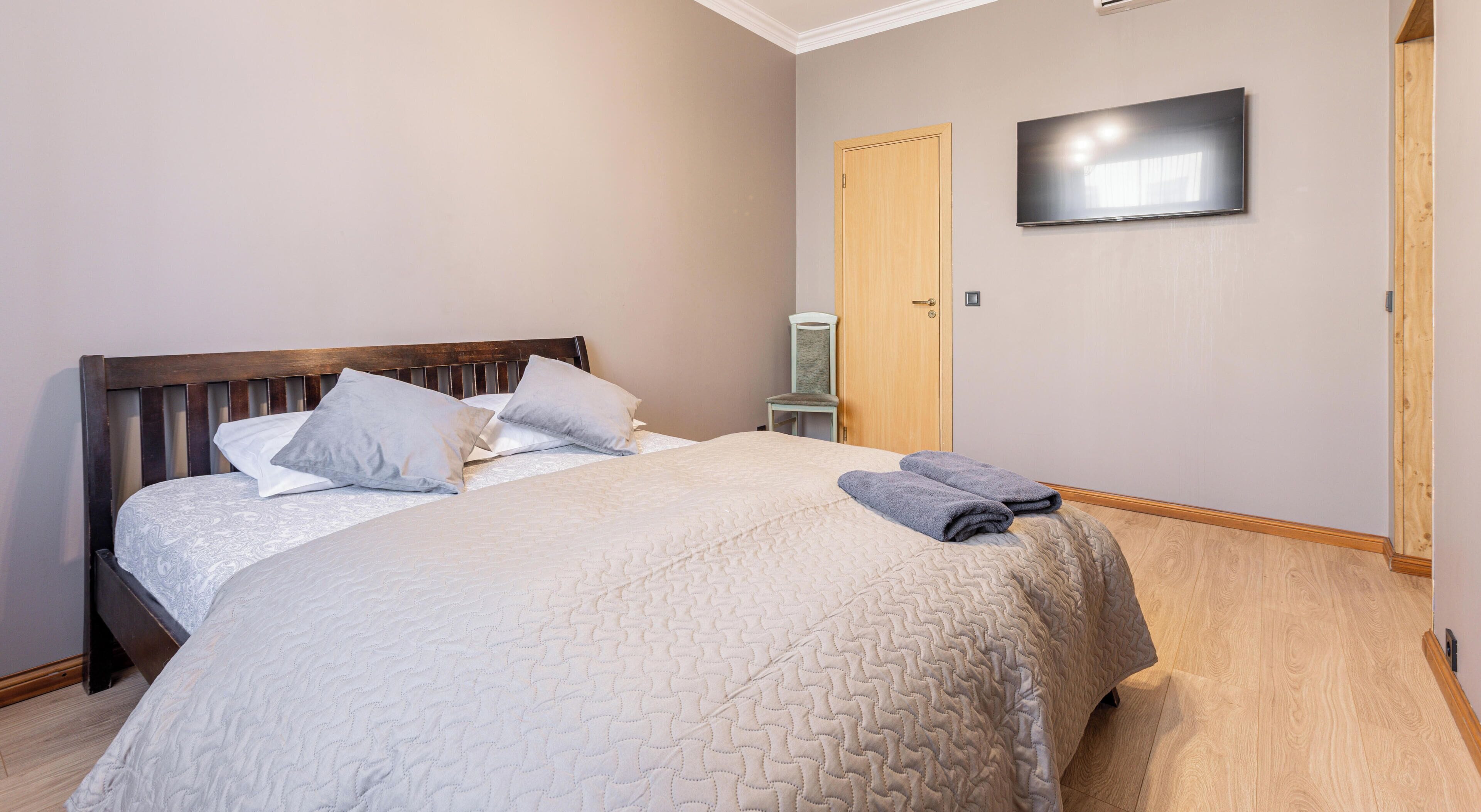 Ein ordentlich bezogenes Bett mit Kopfteil, ein an der Wand befestigter Flachbildfernseher und eine Holztür.