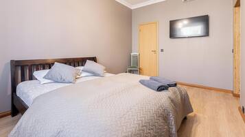 Ein ordentlich bezogenes Bett mit Kopfteil, ein an der Wand befestigter Flachbildfernseher und eine Holztür.