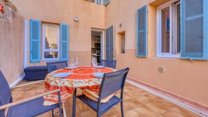 Un área de comedor al aire libre con una mesa preparada para cuatro personas, rodeada de sillas, en un patio empedrado con un edificio al fondo.