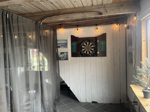 A room with a dartboard mounted on a wooden wall, a window with curtains, and a potted plant on a wooden table. - Cozy Coastal Haven in Havdhem (Havdhem)