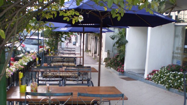 Un café en plein air avec des tables et des chaises sous des parasols bleus, entouré de plantes et de fleurs.