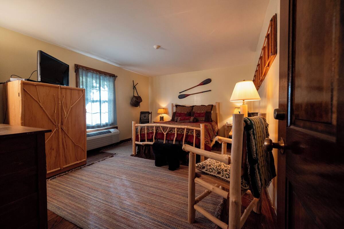 A cozy bedroom with a bed, a wooden chair, a desk, a television, and a window with curtains.