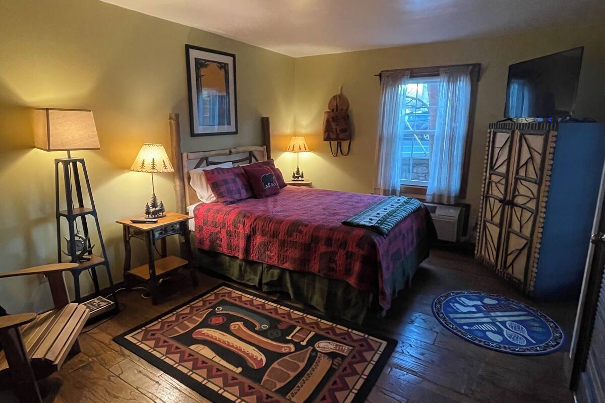A cozy bedroom with a bed, two bedside lamps, a window with curtains, a wooden floor, and a patterned rug.