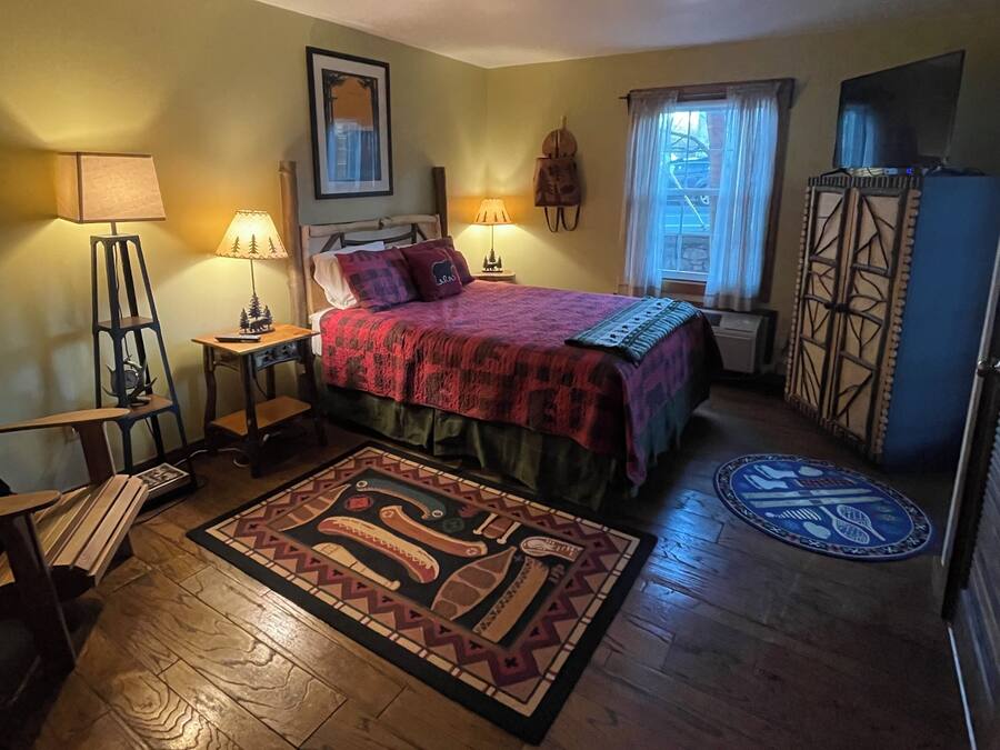 Un dormitorio acogedor con cama, dos lámparas de noche, una ventana con cortinas, suelo de madera y alfombra estampada.
