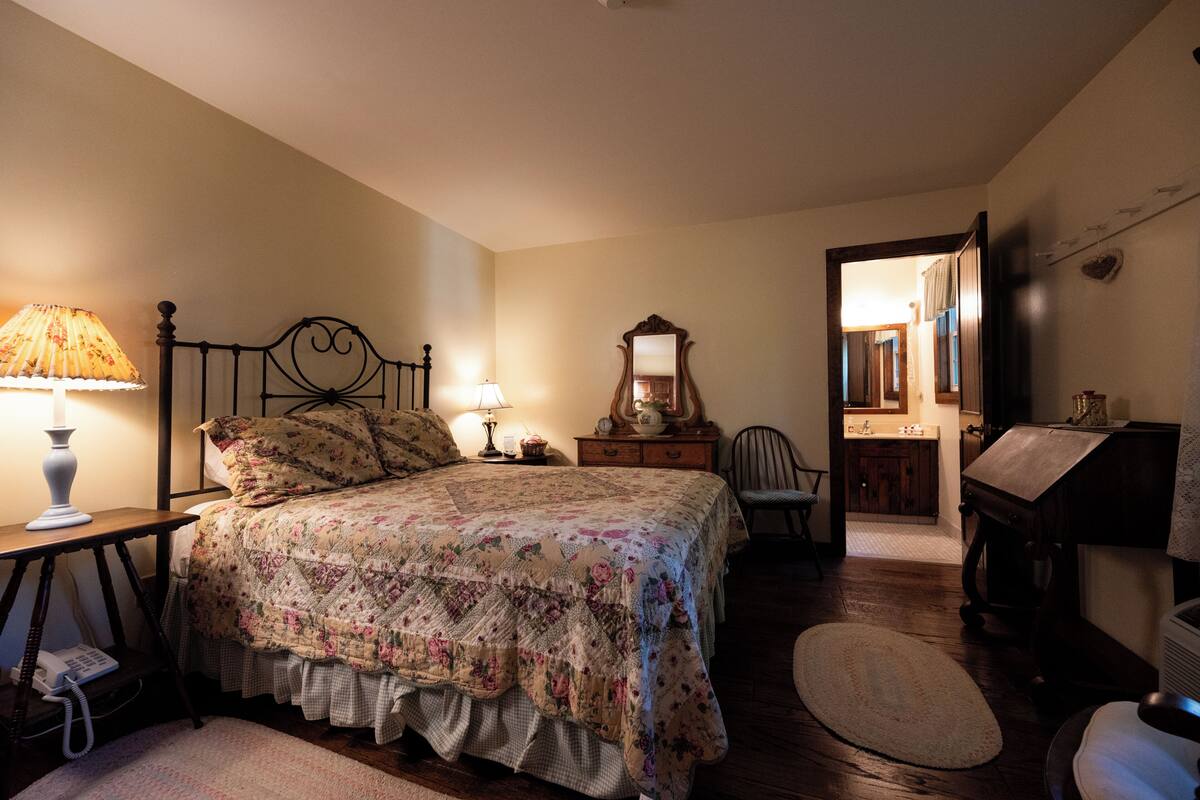 A bedroom with a bed, bedside lamp, dresser, mirror, chair, and a small table with a lamp.