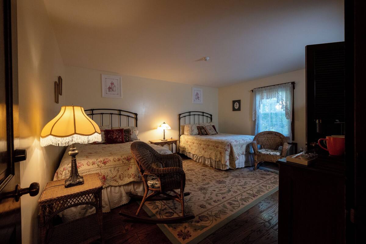 A cozy bedroom with two beds, a rocking chair, a small table with a lamp, and a window with curtains.