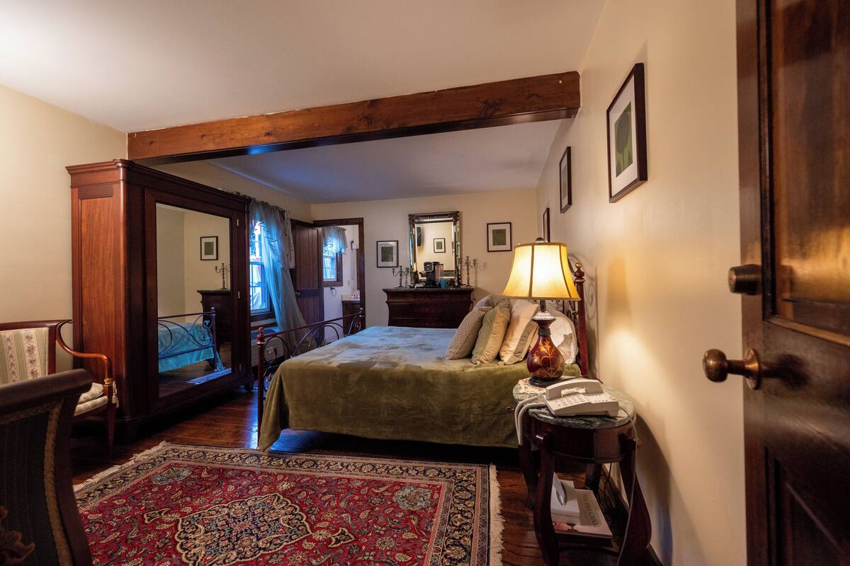 A traditional bedroom with a four-poster bed, a wooden chair, a bedside table with a lamp, and a large mirror on the wall.