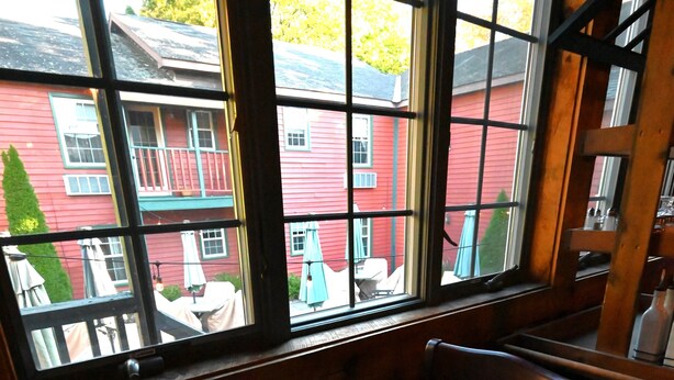 A dining table set for two with red plates, silverware, and a wine glass, viewed through a window overlooking a red building.