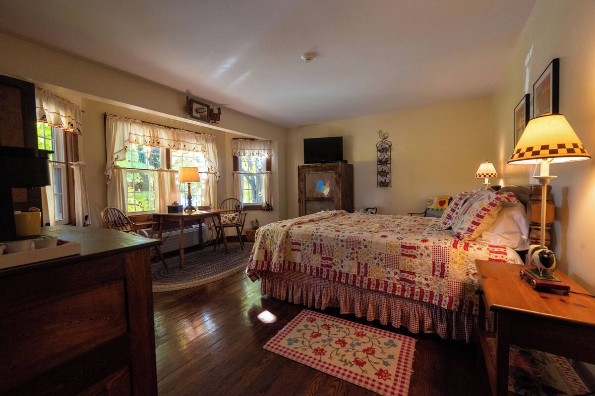 A cozy bedroom with a bed, a desk, and a chair. There is a window with curtains and a lamp on the bedside table.