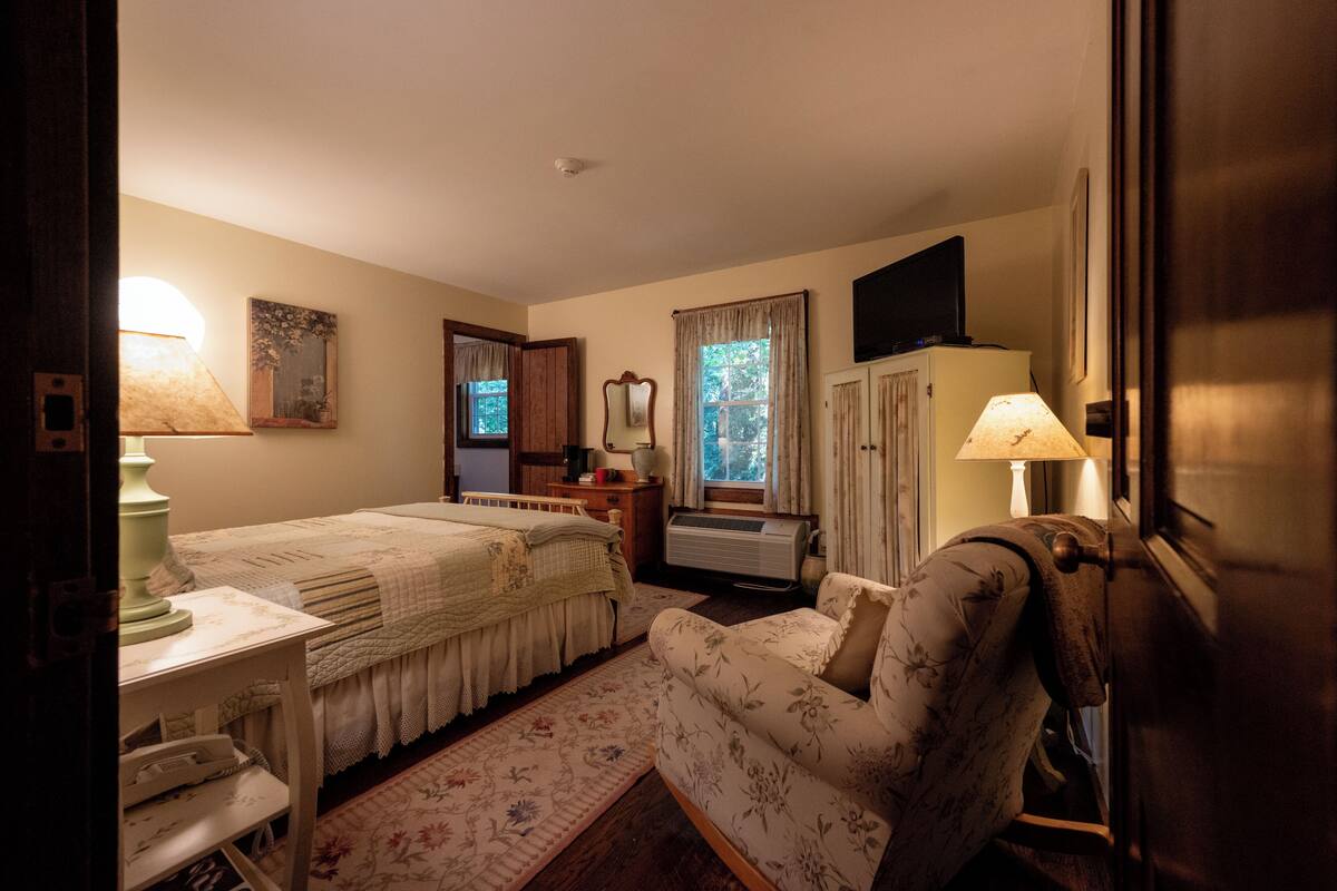 A bedroom with a bed, bedside table, lamp, chair, and a television on the wall.